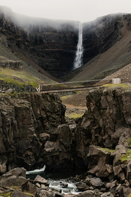 a bridge spanning over water between rocks with a waterfall in the background