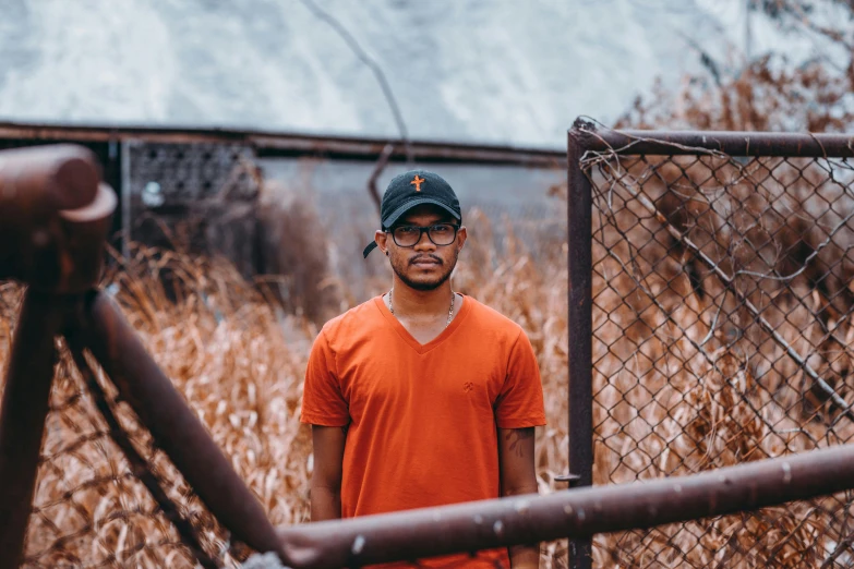 a young man wearing an orange shirt and hat with a rusty fence