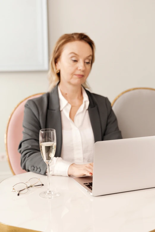 a woman in business attire sits on a table, looking at a laptop computer and drinking a glass of wine