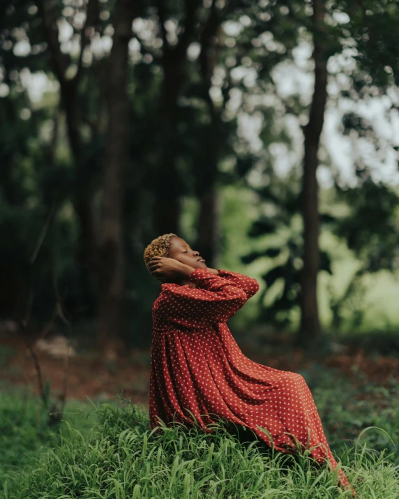 a woman in a red dress and red hair, sitting on the grass near some trees