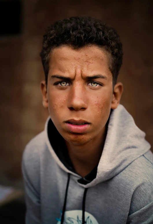 a close - up of a boy in grey and black hoodie with a brick wall and a red building behind him