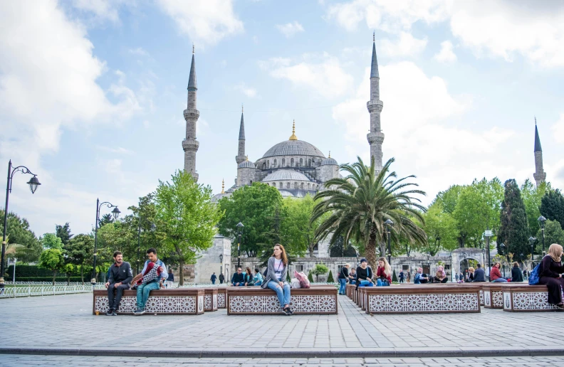 people sit on benches next to a tree and a blue mosque in the background