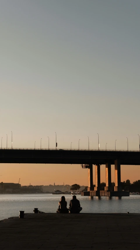 two people sitting under a bridge as the sun sets