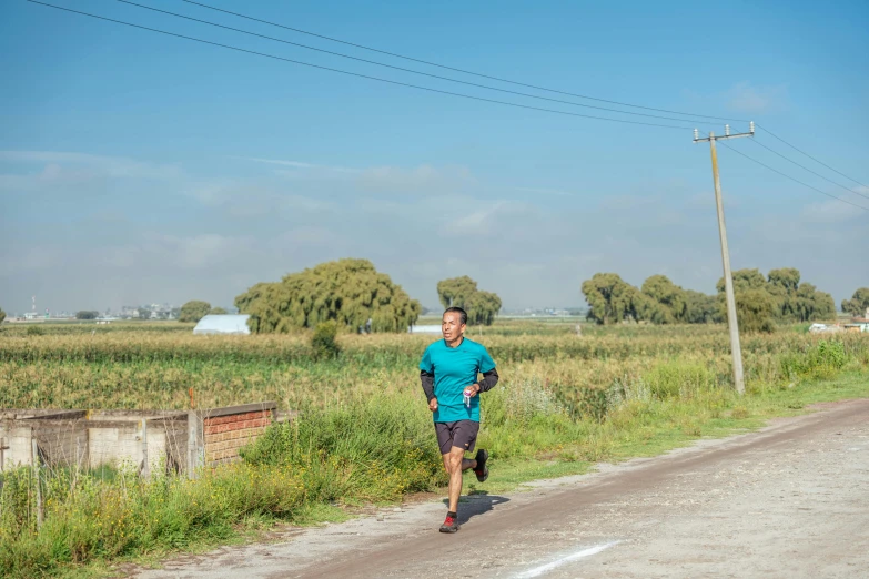 a man in blue shirt running down road next to tall grass and telephone poles