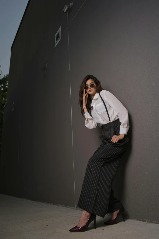 a woman with long dark hair wearing glasses and dress shirt leaning up against a gray wall