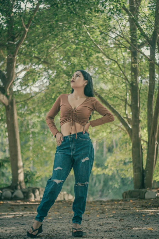 a woman in a brown top and blue jeans stands by a group of trees