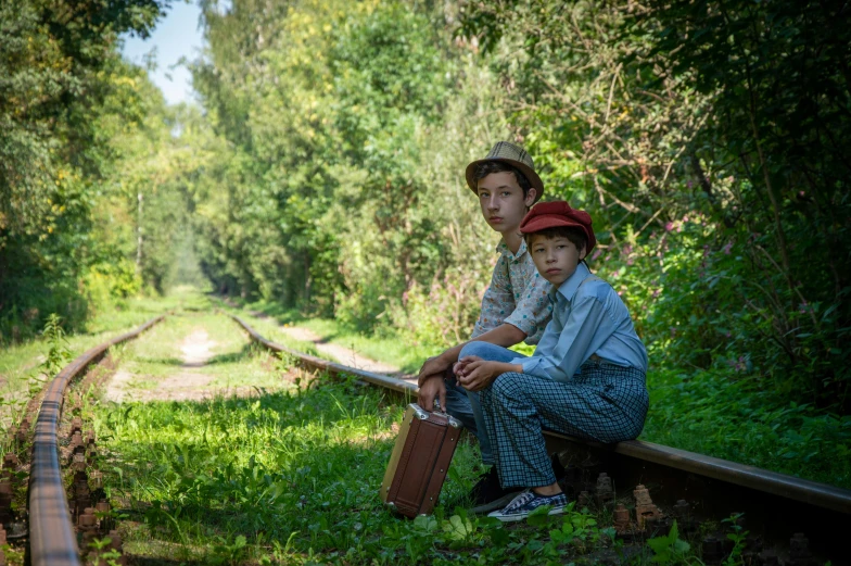 two boys sitting next to each other on the edge of a train track