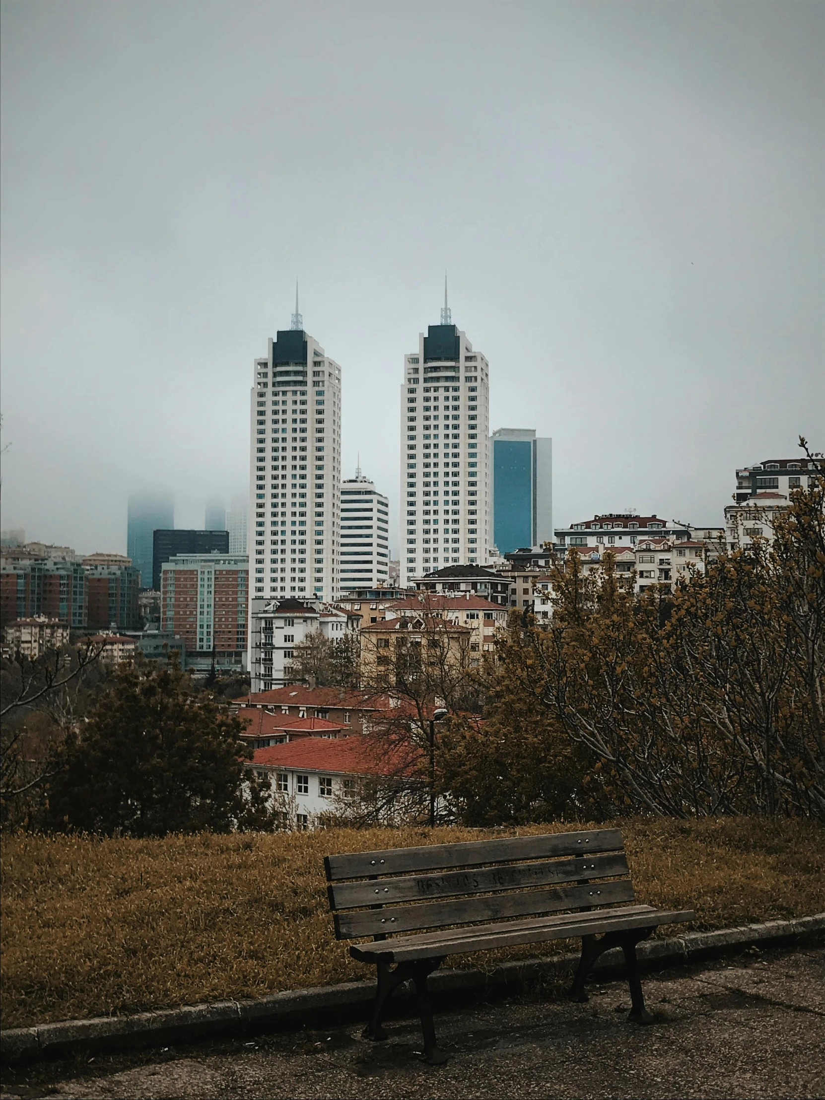 a park bench on a dirt area with city buildings in the background