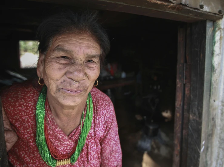 an elderly woman wearing a necklace stands outside a hut