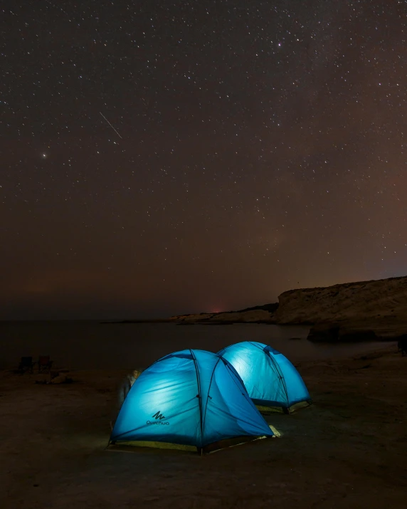 two blue tents with a green top next to the water under a night sky filled with stars