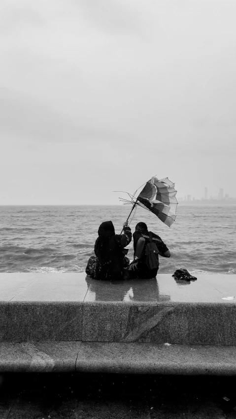 two people sitting on a ledge by the ocean under an umbrella