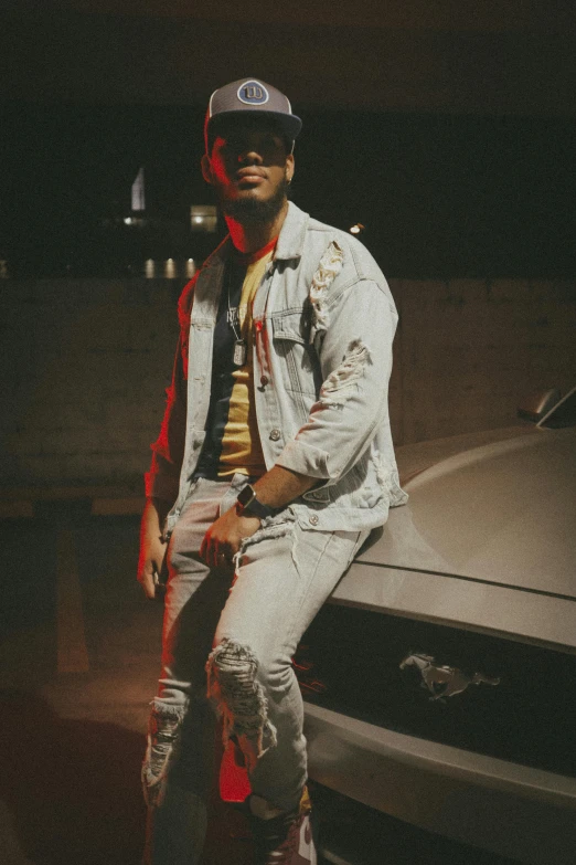 a young man wearing white jeans and a baseball cap sitting on a car