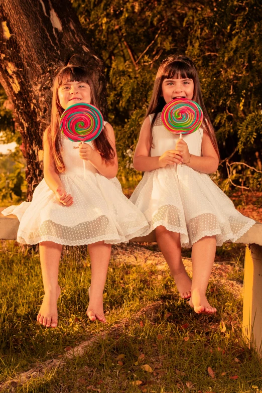 two girls holding circular colorful objects sit on a bench in front of trees