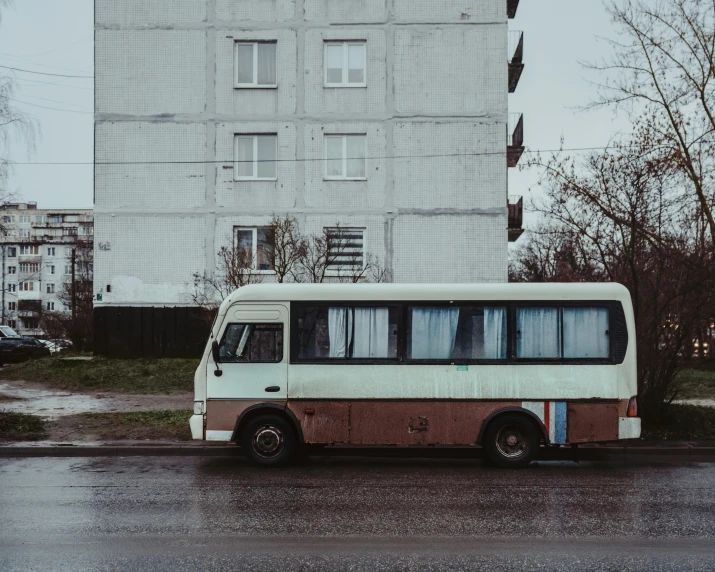 an abandoned white bus sits in front of a grey building on a rainy day