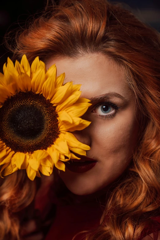 a woman with long curly hair and blue eyes holds a large sunflower in front of her face