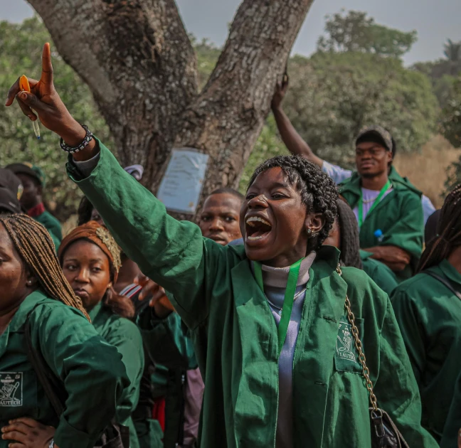 a woman with both hands up, standing in front of several people in green outfits