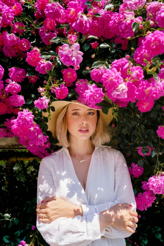 a woman with blonde hair wearing a straw hat and a white shirt in front of a bush of flowers