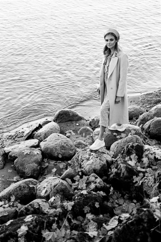 woman in white coat on rocky shoreline by water