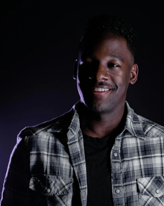 a man in a flannel plaid jacket smiles while standing against a dark background