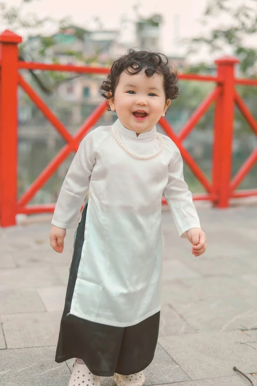 a child with curly hair wearing a white tunic and black pant, holding hands in front of the camera