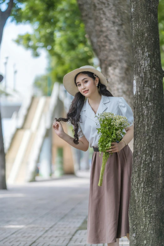 a woman in a hat standing by a tree with a bouquet