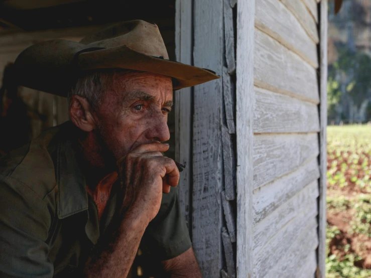 an old man with a cowboy hat leans against the outside of a small house