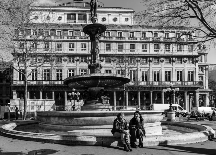 two people sitting on top of a fountain in front of buildings