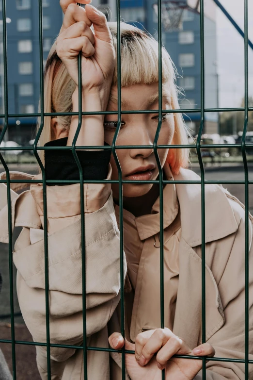 a blonde haired woman with her hair pulled back and hands on her head behind a fence, with a large skyscr in the background