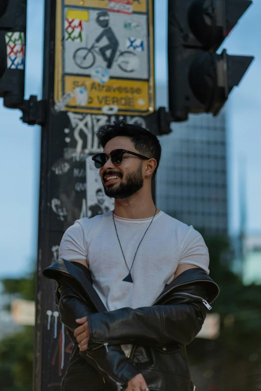 a man smiling while leaning against a pole with a city street sign behind him