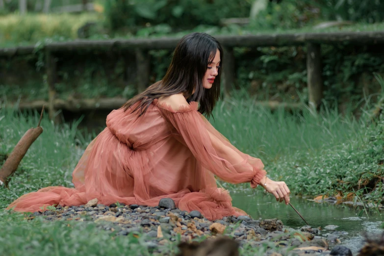 a woman sitting on the ground near a river in a dress