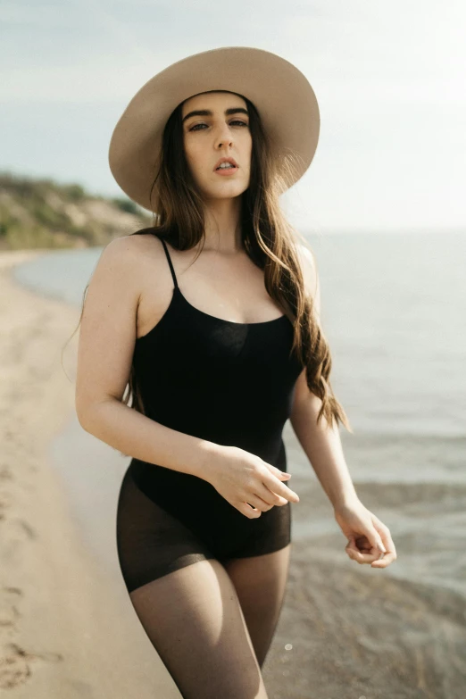 a young lady in a black bodysuit and a hat poses on the beach