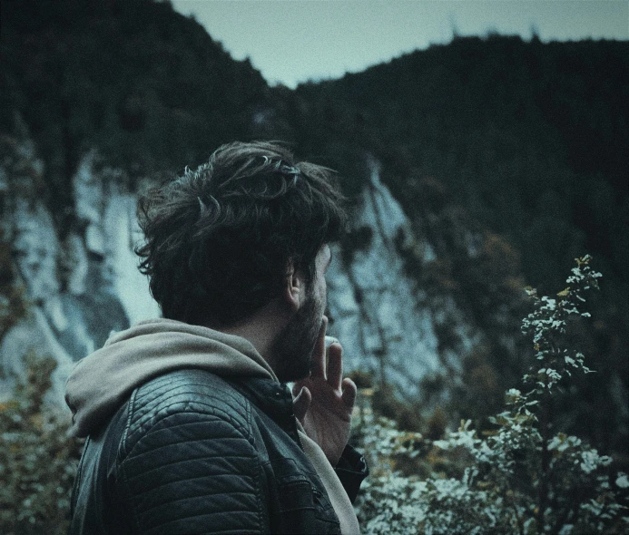 a man stands in the background of mountains while smoking a cigarette