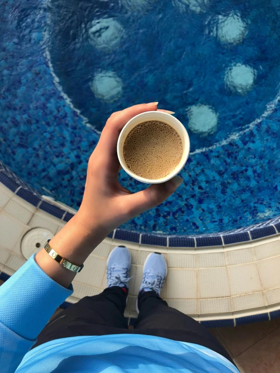 a person with their feet on the edge of a swimming pool holding a coffee cup