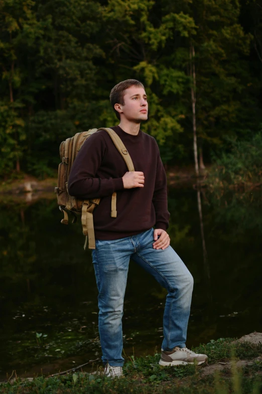 young man wearing blue jeans with backpack leaning on the bank by the river