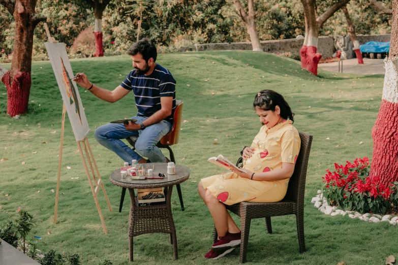 an older lady and man are sitting in chairs in a park, one is painting on canvass while the other sits on a chair