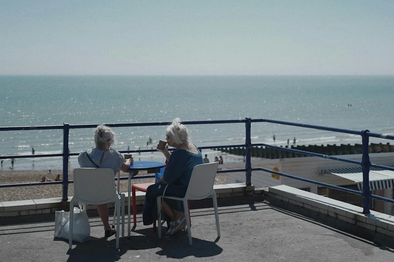 two elderly people sit at an outside dining table by the ocean