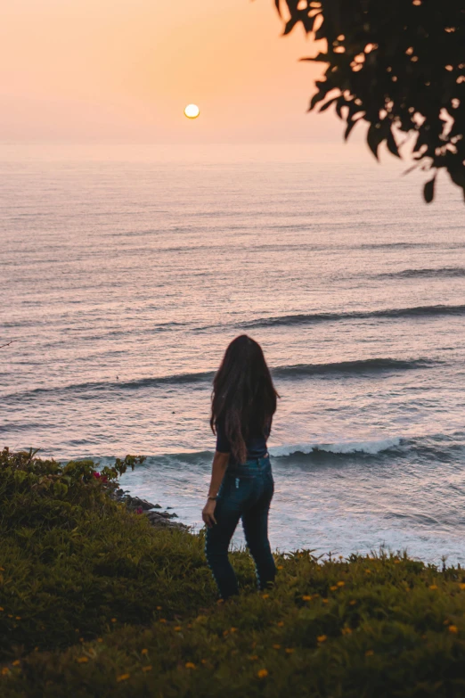 a person in jeans standing on the edge of a cliff looking at the water