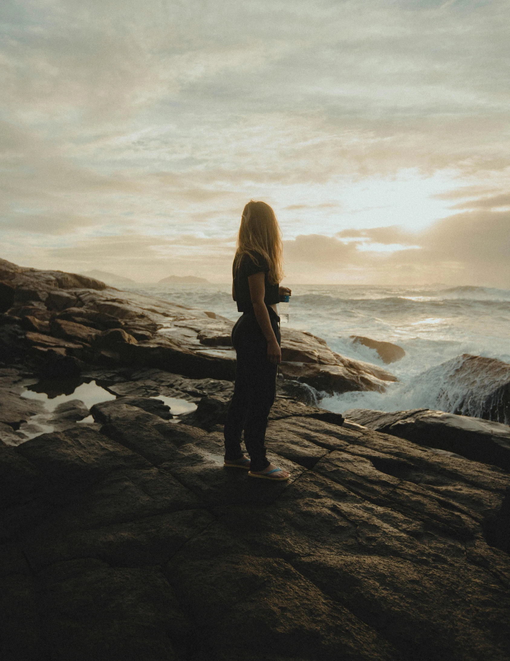 a woman stands at the edge of rocks near the ocean