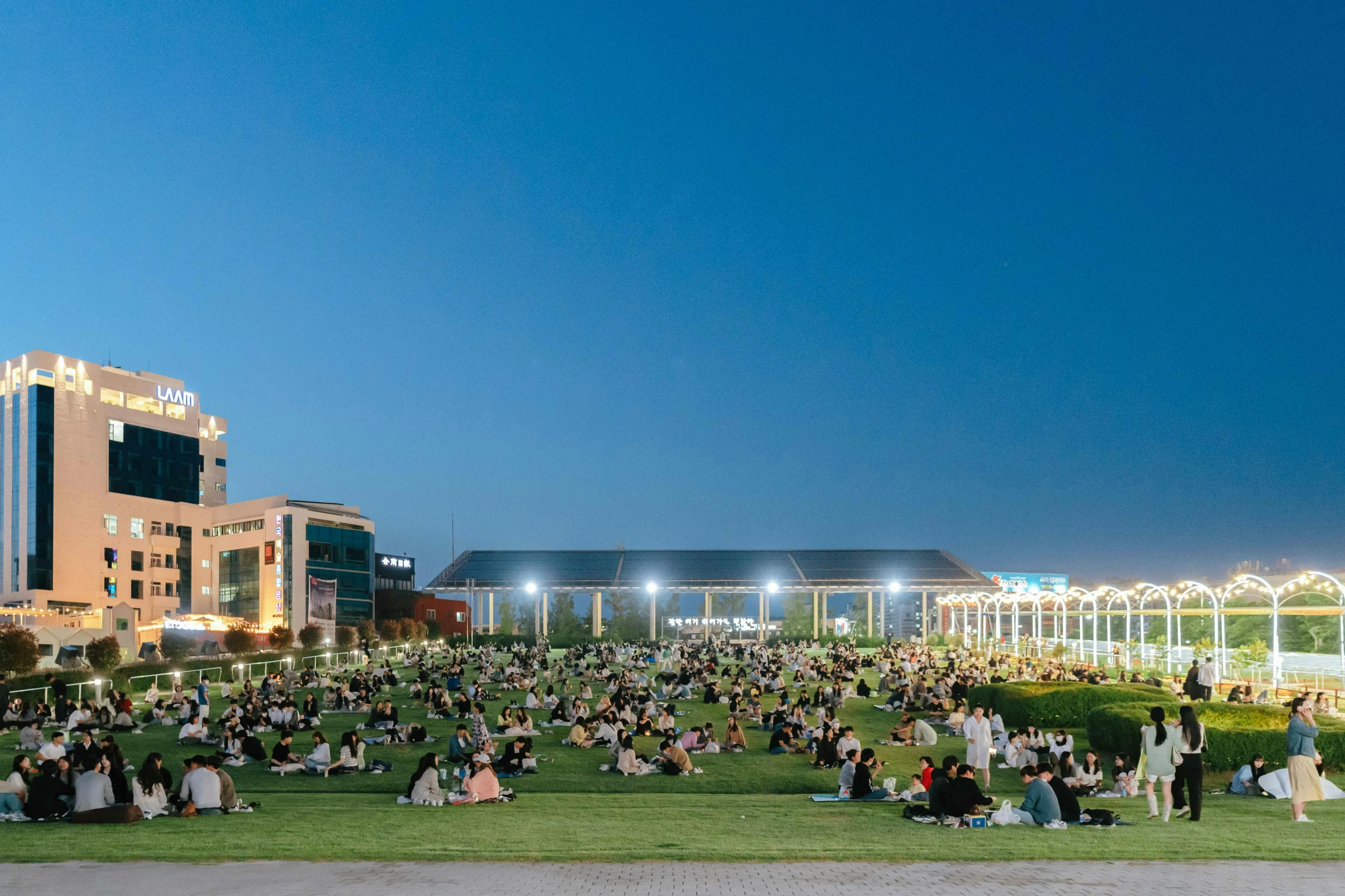 people are gathered in the grass in front of a large building with a lot of lights on it