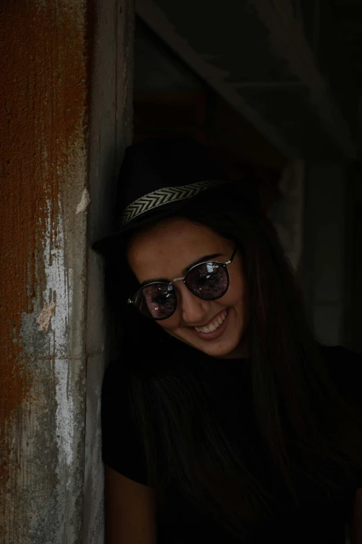 a girl with dark hair in sunglasses and a fedora leaning against the corner of a wooden building