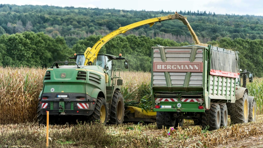 a green machine moving through a large field next to trees