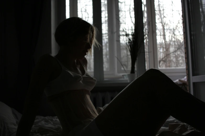 a woman sitting on her bed alone with a window in the background