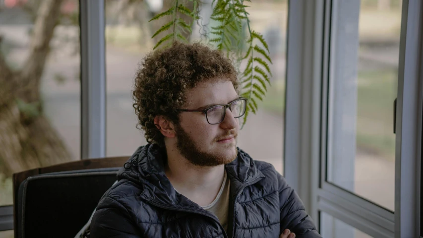 a man with long curly hair sits at a desk near a window