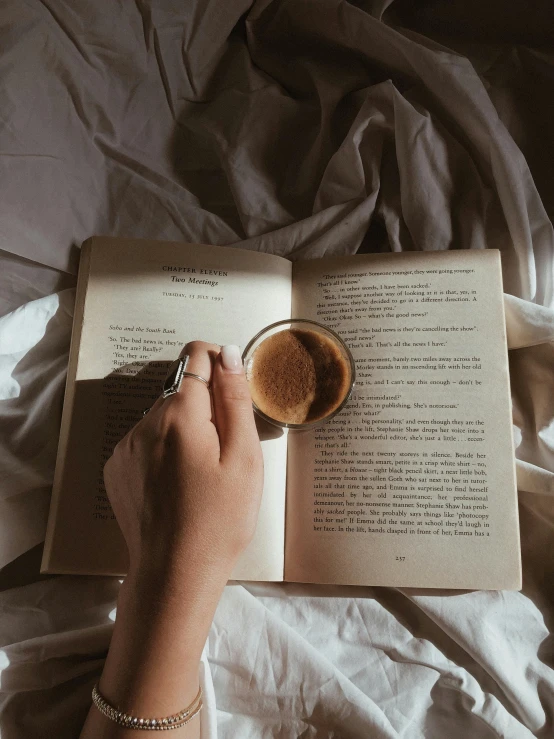 a woman lying in bed while reading a book and drinking coffee