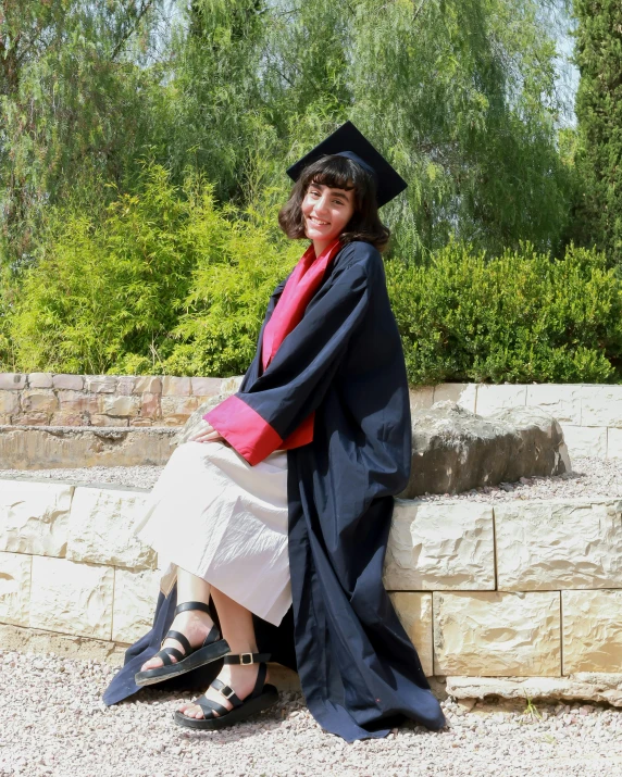 a woman wearing a gown, hood, and graduation cap is sitting on a stone wall