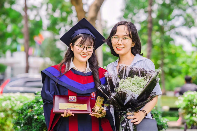 two students in robes and graduation gowns smile as they hold flowers