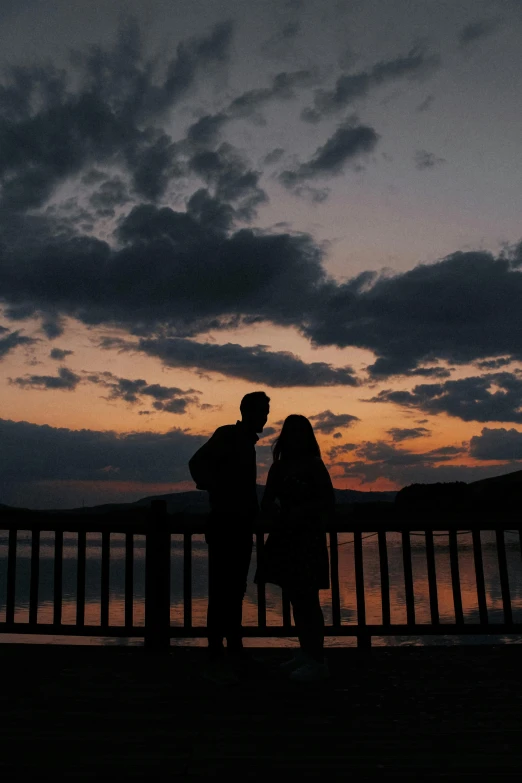 a couple stands against a railing as the sun sets over a body of water