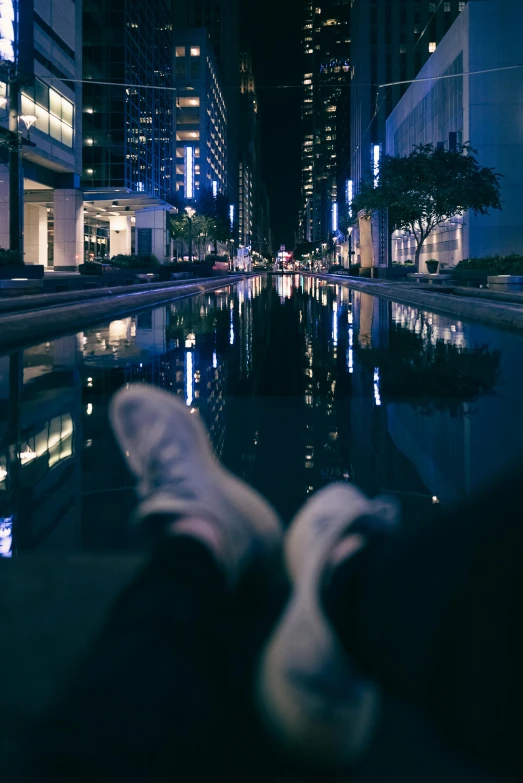 someone's feet sitting on the edge of a waterway in a city at night