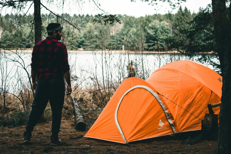 a young man is standing in front of a tent on the shore of a lake