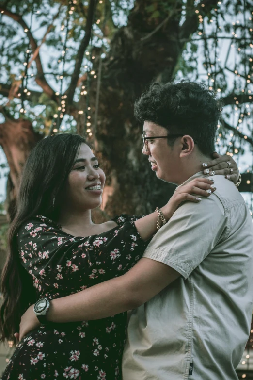 a woman holding a man near trees with lights in the background